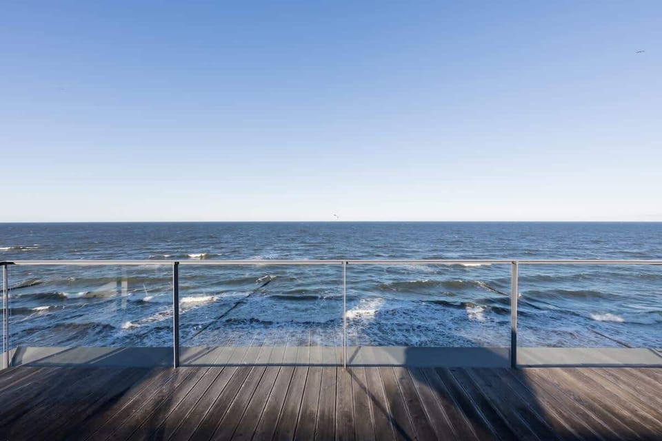 A balcony with a wooden deck and railing, overlooking a calm sea. The horizon stretches far into the distance, creating a peaceful scene.