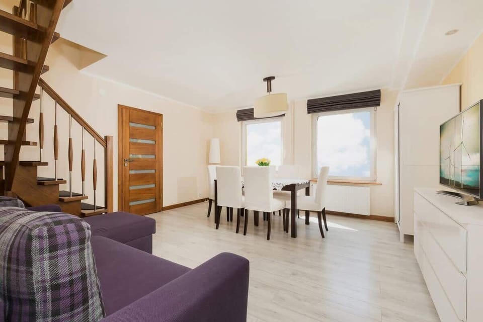 A bright living room with a sofa and table for six, behind which there are large windows that let in plenty of natural light.