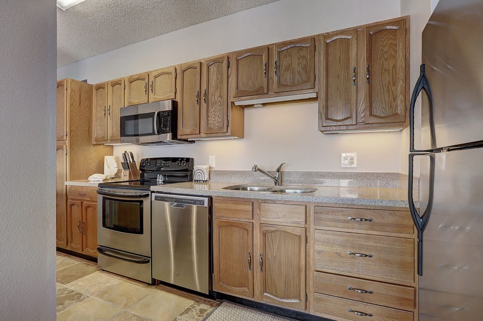 A kitchen featuring wooden cabinets, stainless steel appliances including a stove, microwave, dishwasher, and refrigerator. The countertop includes a sink, knife set, and toaster. Tile flooring is visible.