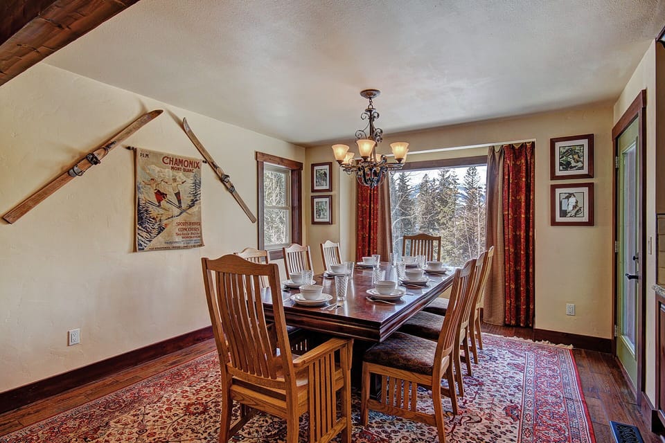 A dining room with a wooden table set for eight, wooden chairs, a chandelier, a large window with curtains, wall art, and a rug on a wooden floor. Two vintage skis are mounted on the wall.