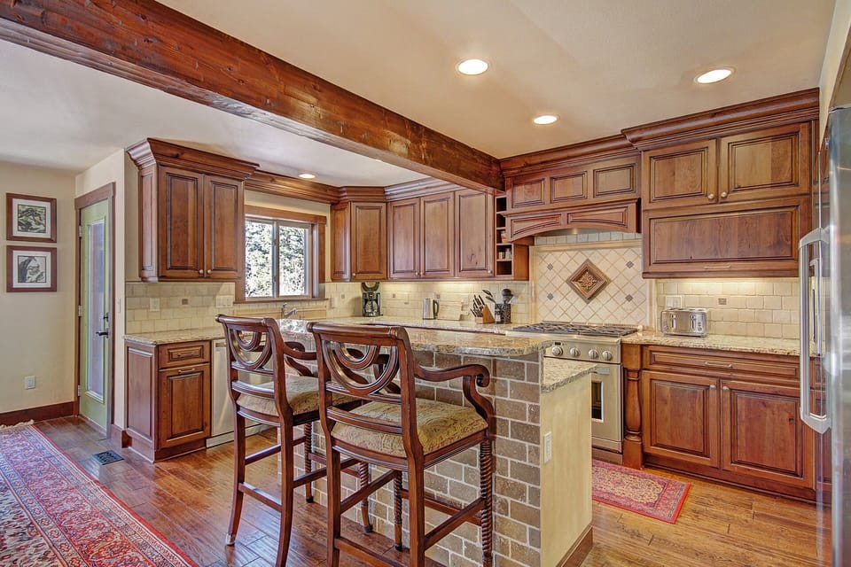 A kitchen interior with wooden cabinets, granite countertops, a central island with two chairs, stainless steel appliances, tiled backsplash, and wood flooring illuminated by recessed lighting.