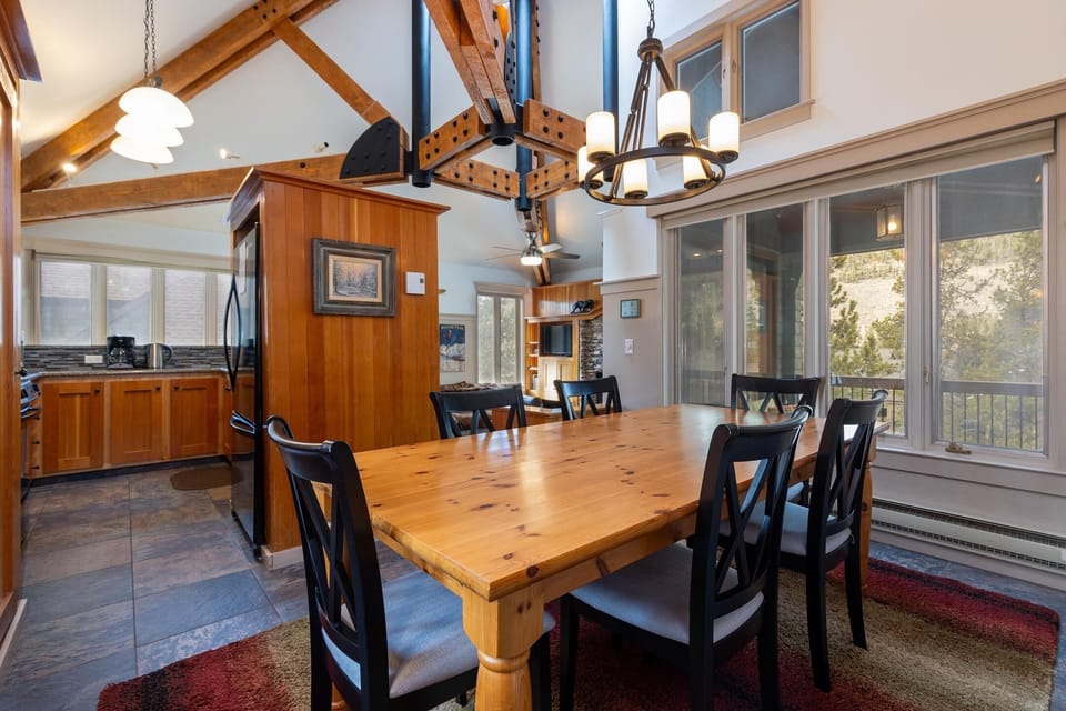 A spacious dining room with a wooden table, six black chairs, large windows, a chandelier, and an attached open kitchen with wooden cabinets.