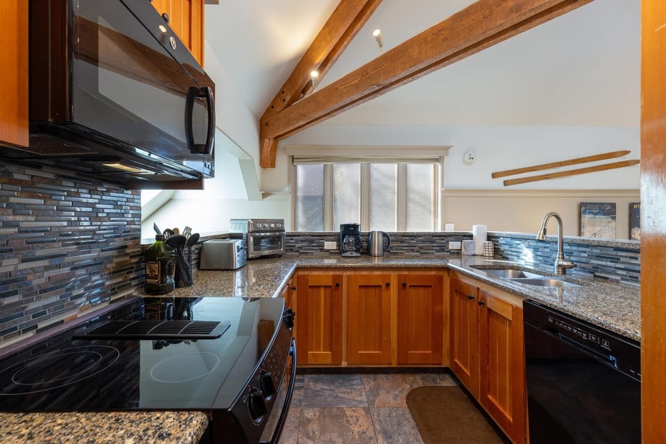 A modern kitchen with wooden cabinets, granite countertops, a black stove, microwave, toaster, coffee maker, and a dishwasher under a large window. The space features exposed wooden beams.