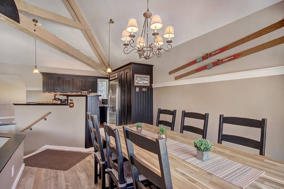 A dining room with a wooden table set for six, black chairs, a chandelier, and rustic ski decor on the wall. The room features wooden beams on the ceiling and a modern kitchen area in the background.