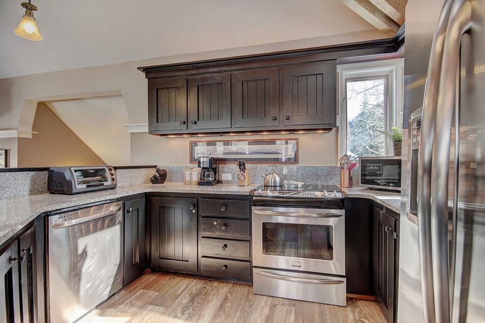 A modern kitchen featuring black cabinets, stainless steel appliances including a dishwasher, an oven, a microwave, and a refrigerator. A window above the counter allows natural light to enter.