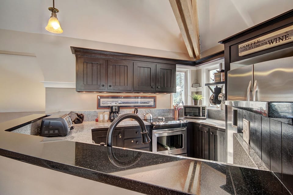Modern kitchen with dark wooden cabinets, granite countertops, stainless steel appliances, a black sink faucet, and a small window. A light fixture hangs from the ceiling and a "Coffee" sign is visible.