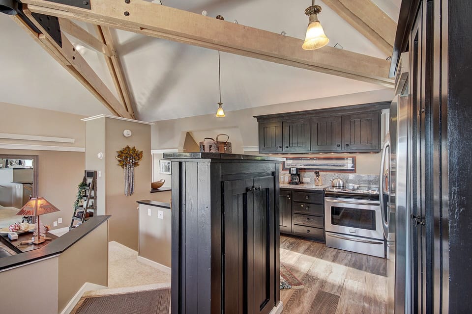 A modern kitchen with wooden beams features dark cabinets, stainless steel appliances, and an island in the center. Pendant lights hang from the ceiling, and a staircase is seen in the background.