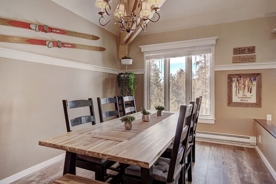 A dining room with a wooden table and chairs, decorated with skis on the wall. A window with a nature view is in the background. A potted plant and wall decor add to the cozy atmosphere.