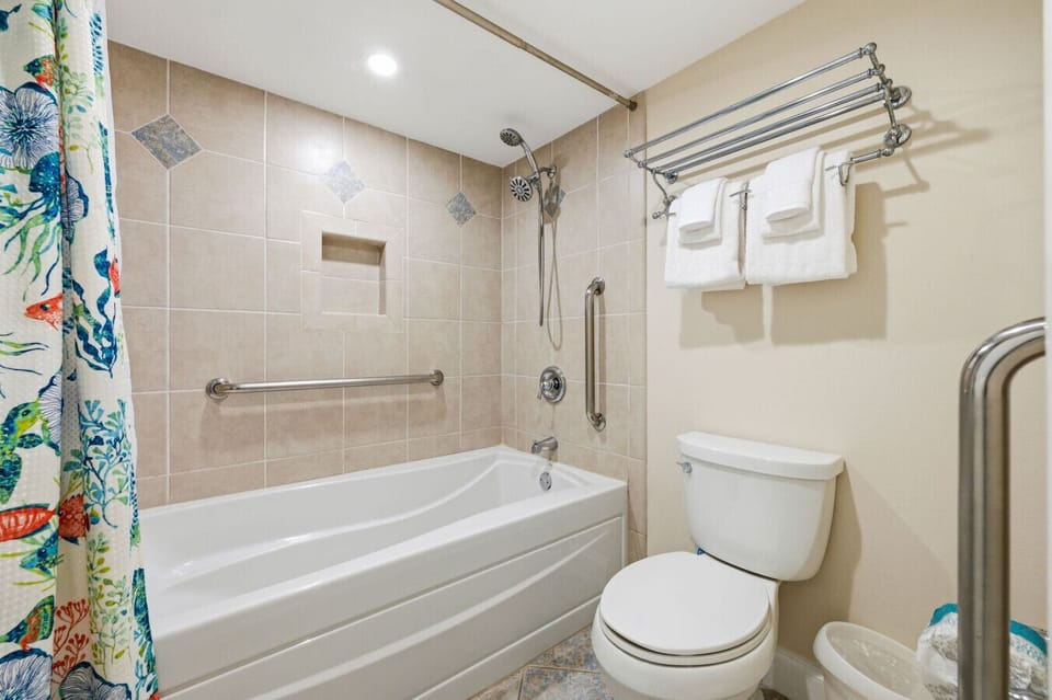 Coastal Vacation Condo Primary Bathroom Featuring a Bathtub and Shower Combination, Tiled Surround, Convenient Grab Bars, Handheld Showerhead, and Fresh Towels