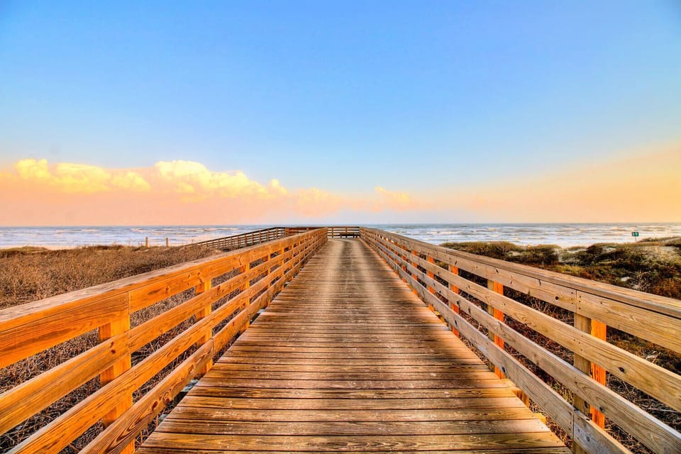 A scenic wooden boardwalk stretches across coastal landscape toward the beach, with warm sunset colors painting the sky above natural surroundings.