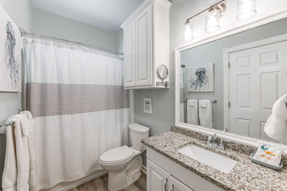 Your private bathroom features granite countertops, white cabinetry, and coastal artwork creating a fresh, spa-like retreat.