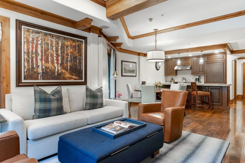 Living area with cream sofa, plaid pillows, leather chairs, blue ottoman, and wood-trimmed ceiling beams.