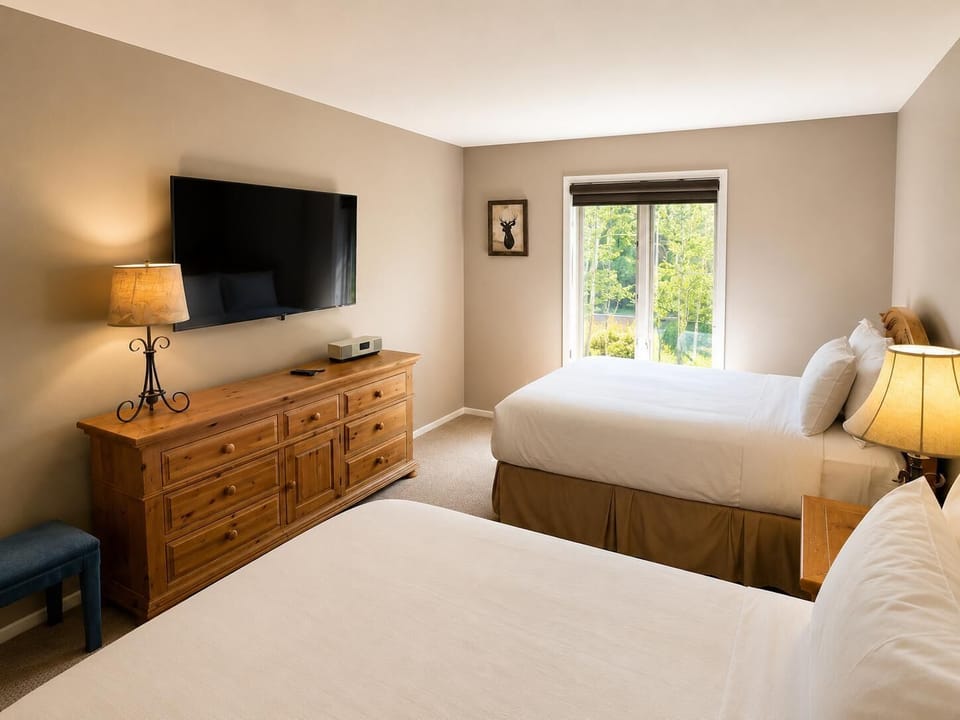 Guest bedroom with two queen beds, wooden dresser beneath wall-mounted TV, and window overlooking greenery.