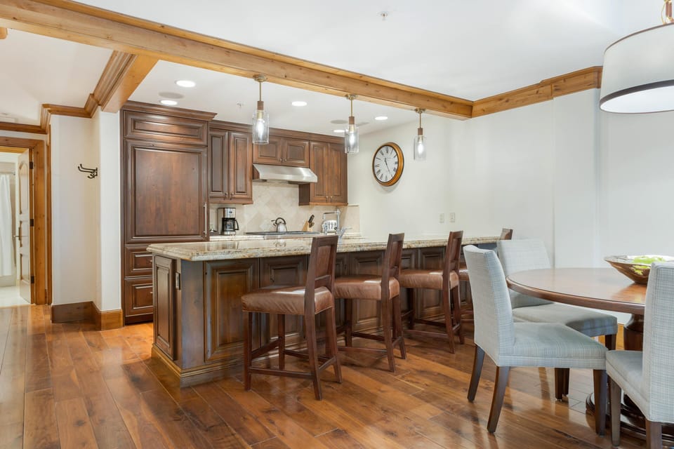 Dark wood cabinetry and leather bar stools line the granite kitchen island beneath pendant lighting.