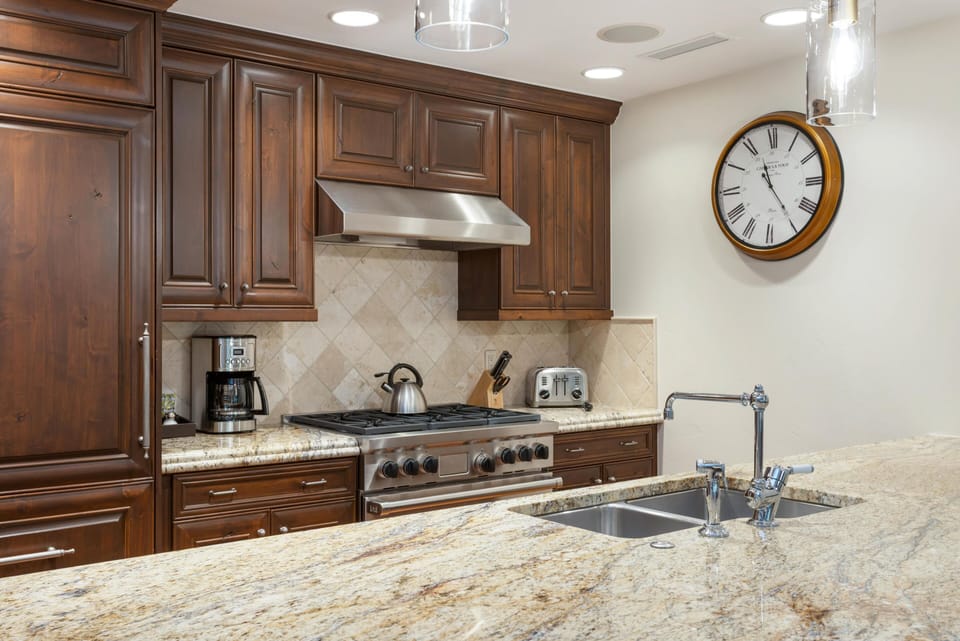 Stainless steel gas range beneath a vent hood, surrounded by dark wood cabinets and beige tiled backsplash.