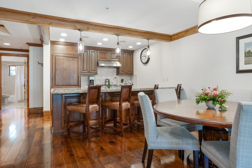 Hardwood floors reflect the overhead lights above a kitchen bar with brown leather stools and a round dining table.
