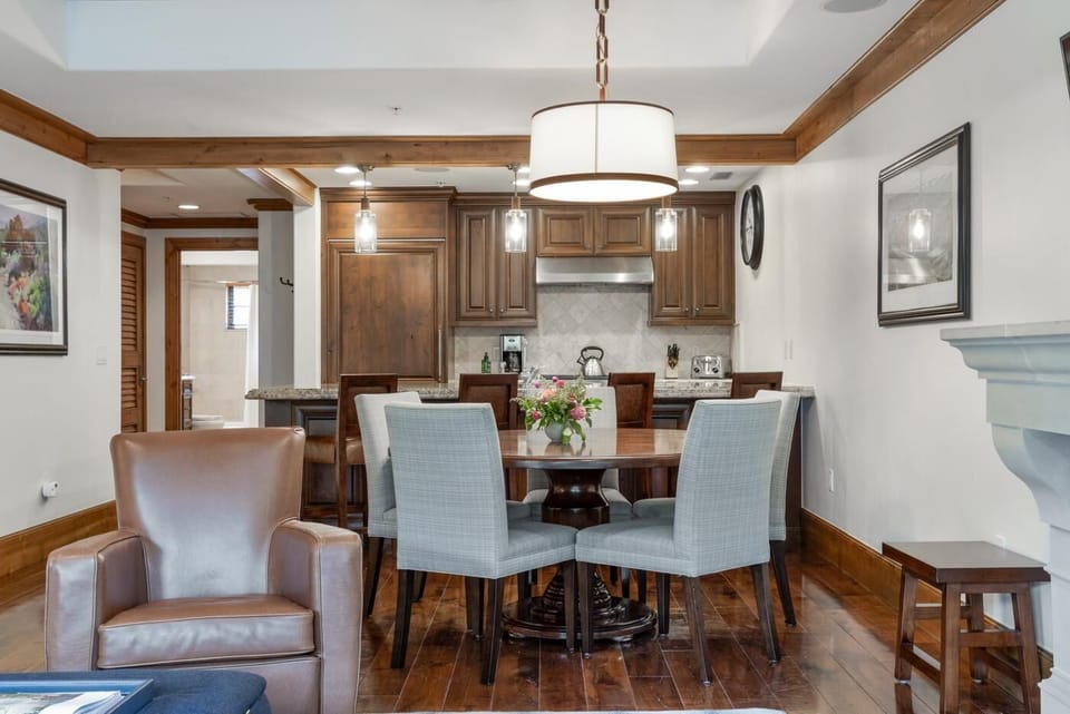 Round wood dining table surrounded by six gray upholstered chairs beneath a drum pendant light.