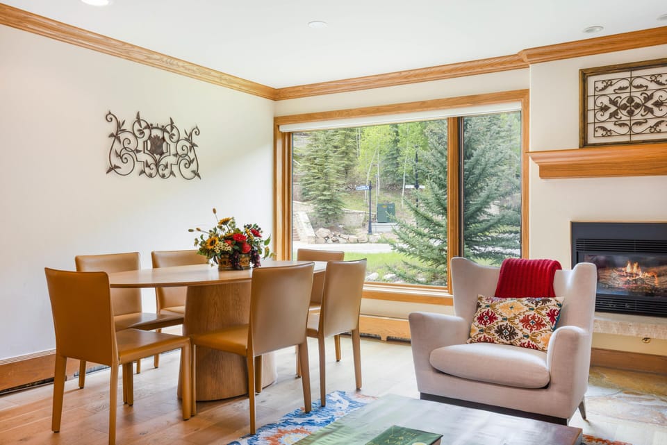 Dining area with tan leather chairs around a wooden table, next to a large window overlooking evergreens.