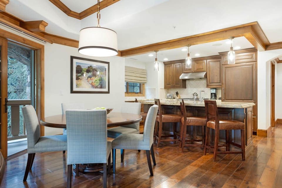 Round wood dining table with upholstered chairs sits beside sliding glass door to balcony. Hardwood floors extend to kitchen.