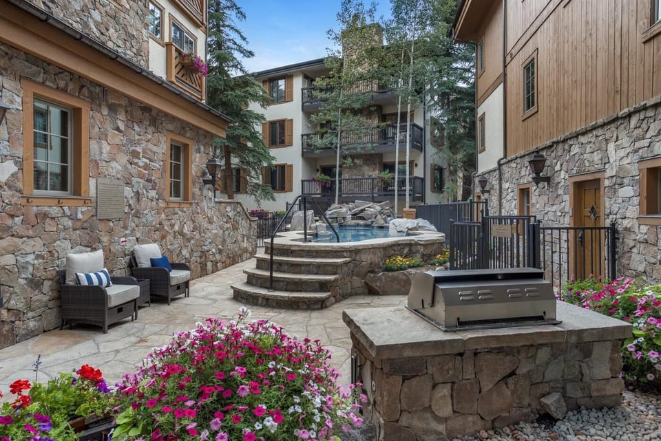 Stone patio with cushioned wicker chairs, built-in barbecue, and a raised hot tub surrounded by flowers.