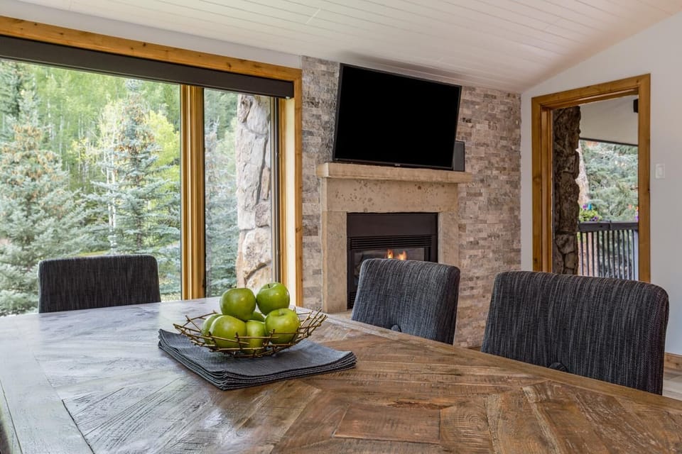Dining table with dark textured chairs overlooks pine trees through large windows near a stone fireplace.