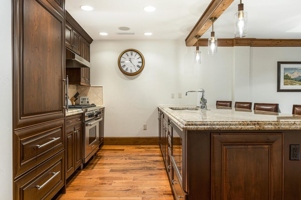 Granite-topped kitchen island with built-in sink and pendant lights contrasts with dark wood cabinetry and warm hardwood floors.
