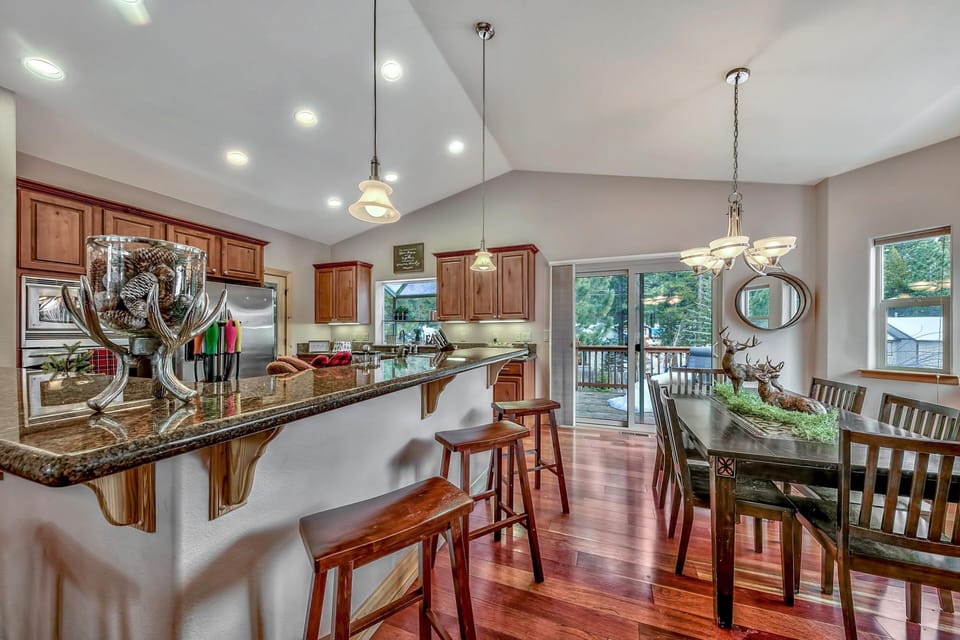 Granite breakfast bar with wooden stools faces kitchen cabinetry and stainless steel appliances under recessed lighting.
