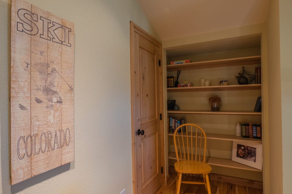 A small room with a wooden door and built-in shelves. On the shelves are books, small decorative items, and a framed photo. A wooden chair is placed in front of the shelves. A "Ski Colorado" sign hangs on the wall.