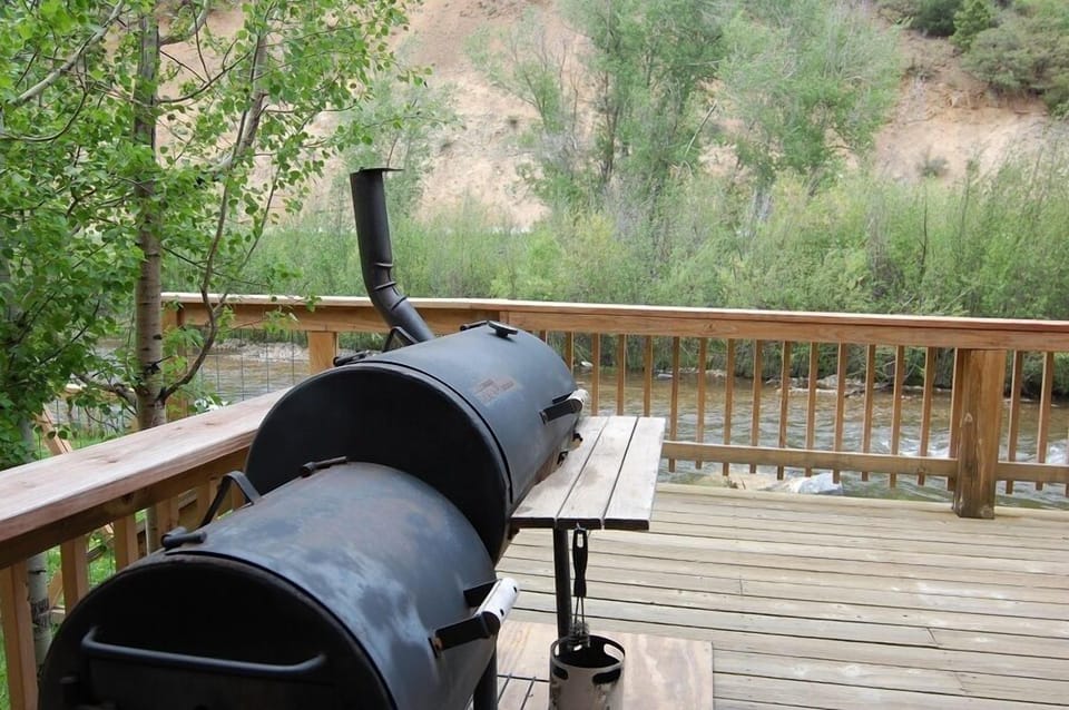 Spacious deck overlooks river and natural hillside landscape. Features a large charcoal smoker-grill ready for outdoor cooking and entertaining. Property is surrounded by desert-mountain terrain.