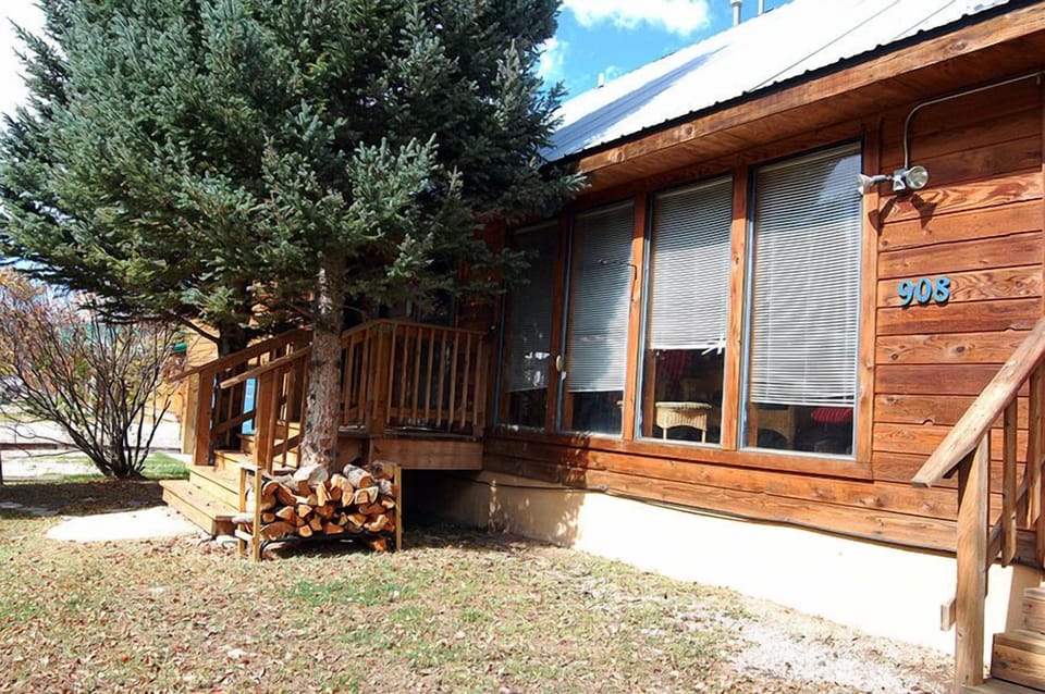 Charming cedar cabin nestled among tall pines with a welcoming front deck and firewood stacked for cozy evenings, offering a serene mountain escape under bright blue skies.