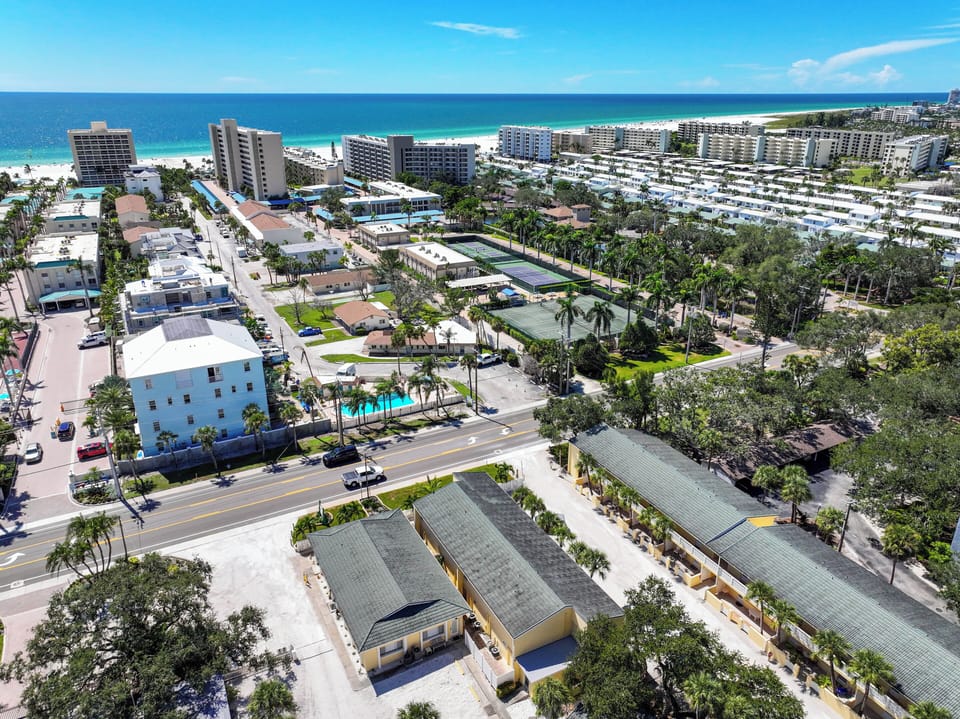 An aerial view showcasing a vibrant beachfront community with pristine white sands, turquoise waters, and the property nestled among palm-lined streets near tennis courts and resort amenities.