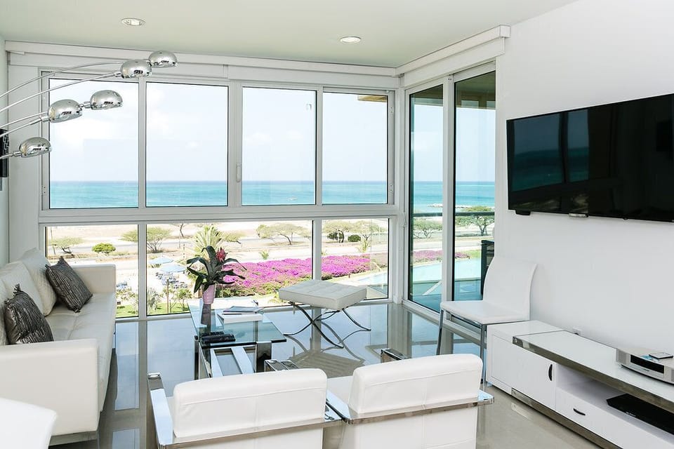 Relax in your bright living room with breathtaking ocean views. Floor-to-ceiling windows frame colorful gardens and sparkling turquoise waters, while modern furnishings create a serene, comfortable space for your coastal retreat.