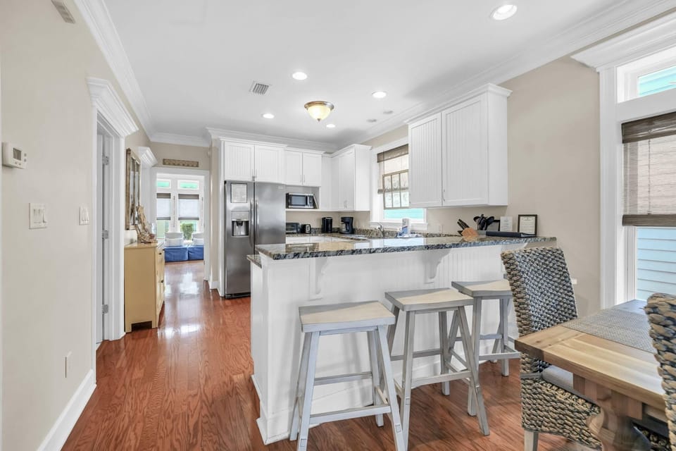 Granite countertop breakfast bar with three wooden stools overlooks white cabinetry and stainless steel appliances.