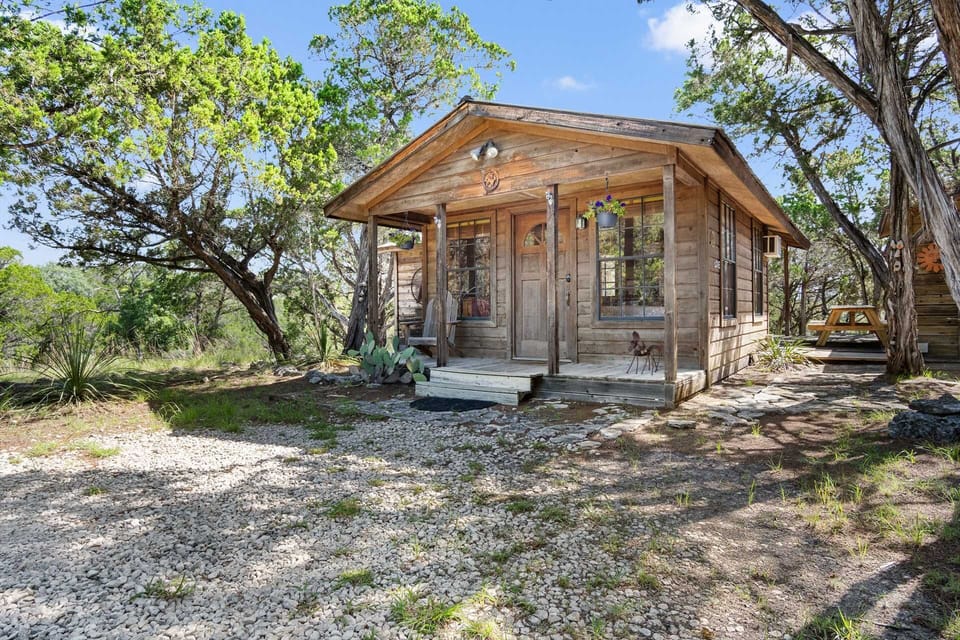 A quaint wooden cabin, nestled among trees, features a welcoming porch with steps, surrounded by gravel and natural vegetation.
