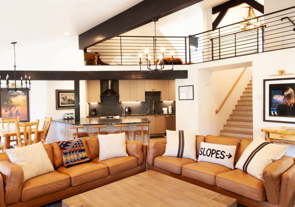 Unwind in your spacious, light-filled living area featuring soaring ceilings with exposed beams, cozy leather sofas, and a fully-equipped modern kitchen—perfect for gathering after a day on the slopes.