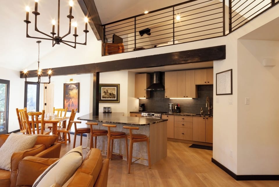 Imagine your mornings here: sunlight spills across warm wood floors in this open living space where every detail—from the vaulted ceiling with exposed beams to the sleek kitchen island—invites you to relax and gather.