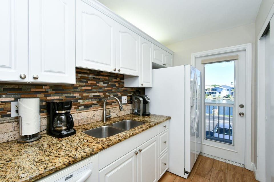 Modern, completely renovated kitchen with granite counter tops, glass tile back splash, opens to living/dining area and window/door overlooking the bay and boat docks