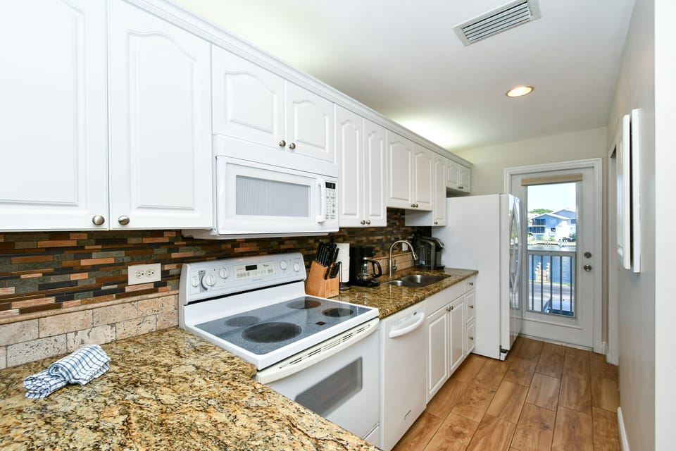 Modern, completely renovated kitchen with granite counter tops, glass tile back splash, opens to living/dining area and window/door overlooking the bay and boat docks