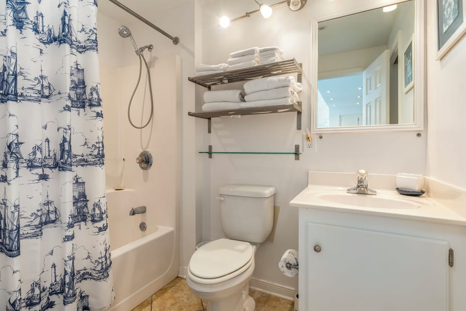 Your bright bathroom features a tub-shower combo with nautical-themed curtain and fresh towels neatly organized on open shelving.