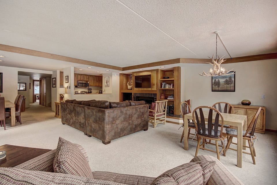 A spacious living room with a brown sectional sofa, a dining table with wooden chairs, a kitchen in the background, and a chandelier made of antlers. The room features wooden accents and neutral tones.