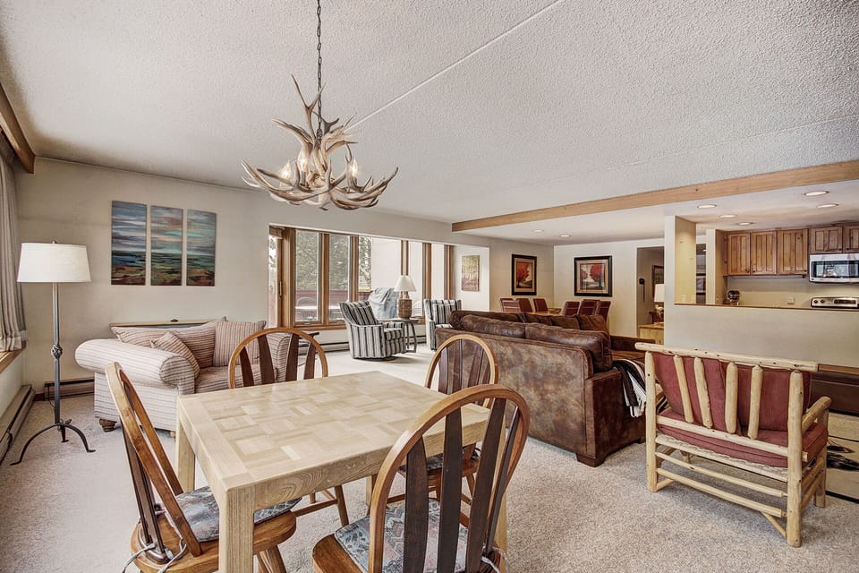 Open living and dining area with wooden furniture, antler chandelier, beige carpet, modern paintings on walls, and window view. Kitchen visible in the background.