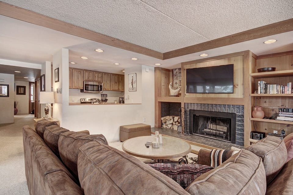 A cozy living room with a large sectional sofa, a circular coffee table, a fireplace with a TV above it, and built-in wooden shelves. An open kitchen is visible in the background.