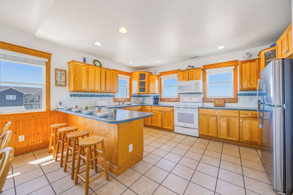 Your spacious kitchen invites shared meals with warm wood cabinetry, stainless appliances, a large island with seating, and abundant natural light streaming through multiple windows.