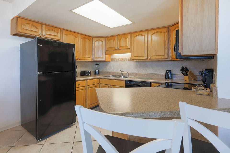 This kitchen features black appliances, wooden cabinetry, and a breakfast bar with white chairs for casual dining