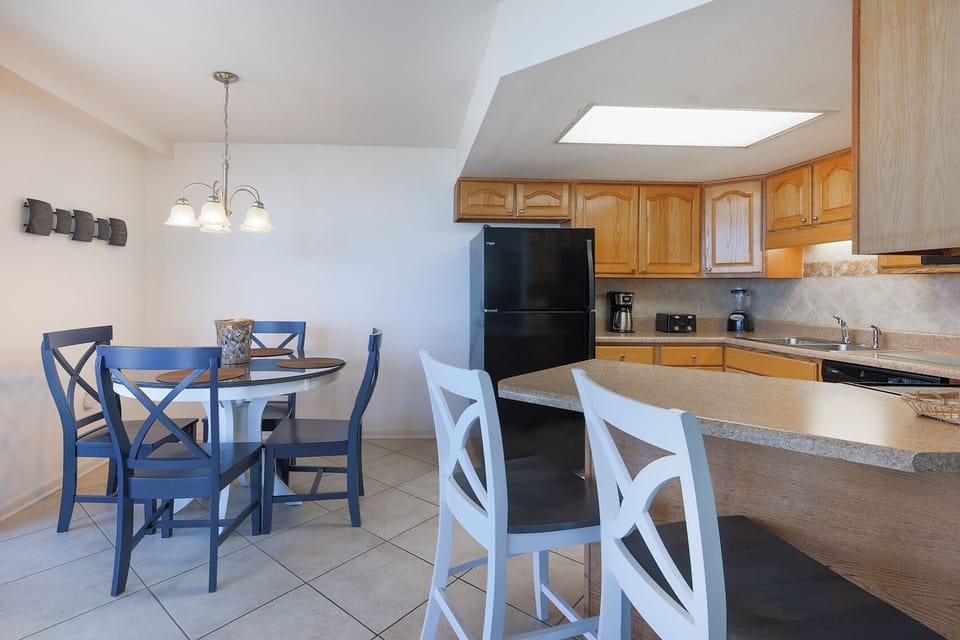 The kitchen features light wood cabinetry, a black refrigerator, and the dining area with a round table and blue chairs