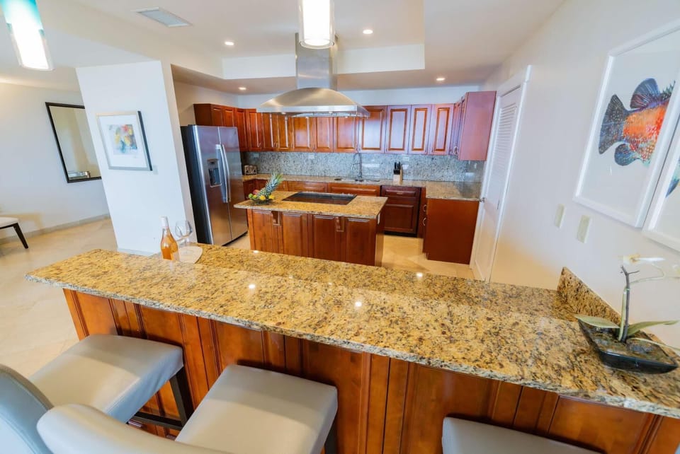 Granite breakfast bar with three cushioned stools overlooks cherry wood cabinetry and stainless steel appliances.