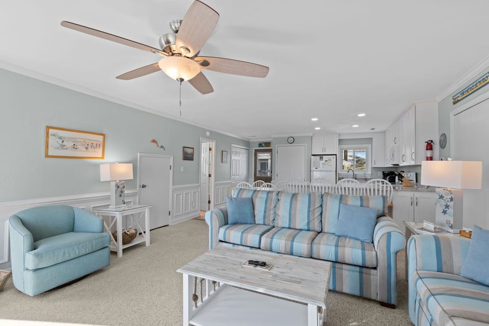 Relax in your coastal-themed living space with comfortable striped seating, gentle ceiling fan breeze, and an open layout connecting to your fully-equipped kitchen.