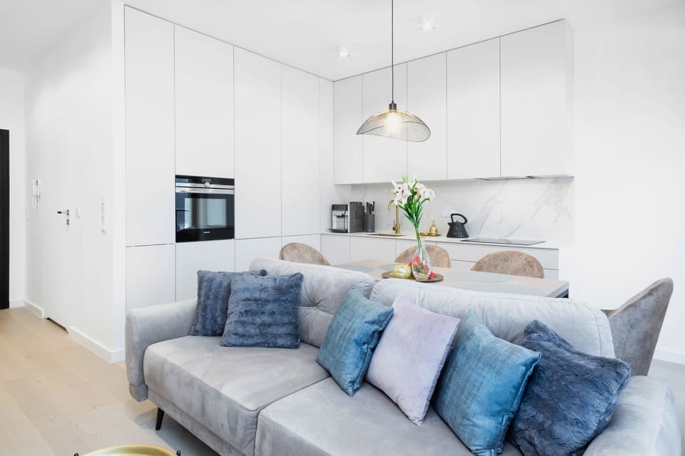 A comfortable grey sofa with decorative cushions behind which you can see the kitchenette with white cabinets and quality appliances. 
