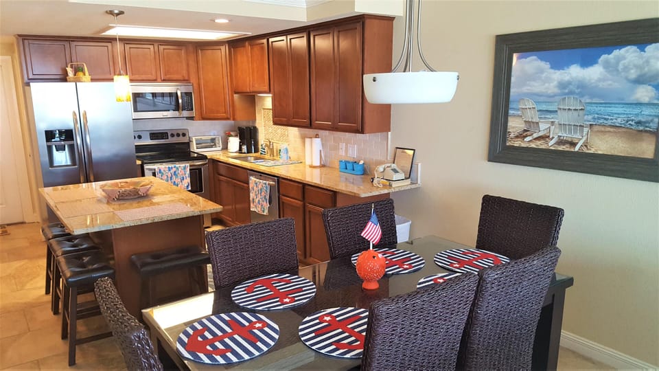 Glass dining table set with woven chairs and nautical placemats beside a granite-topped kitchen island.