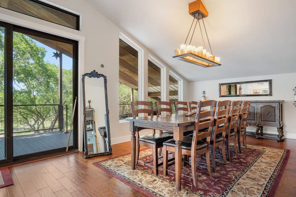 A rustic dining room with a large wooden table and chairs, illuminated by a rectangular chandelier, opens to a scenic balcony.